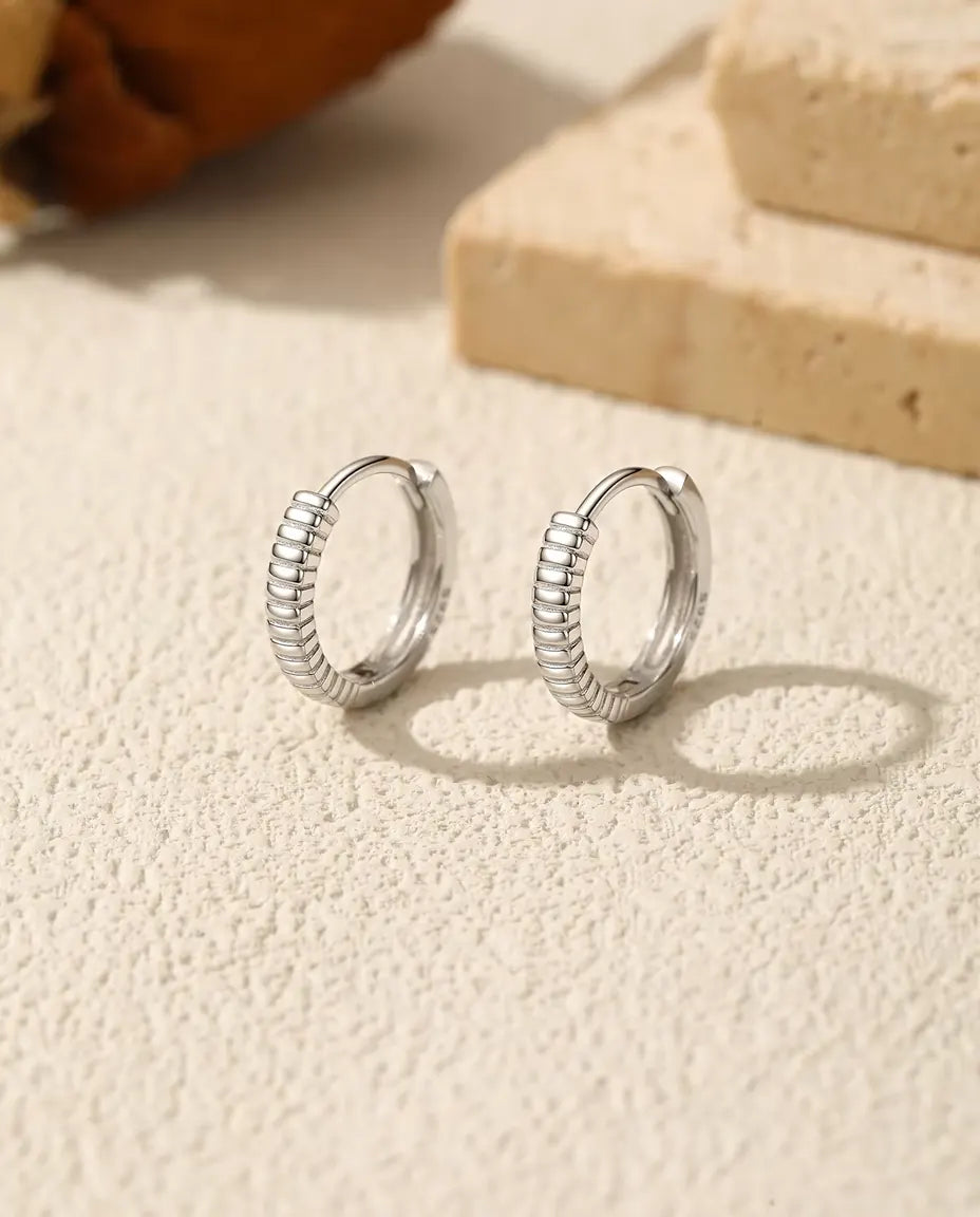Pendientes Aro Huggie Estriados en Plata 925 - el par uno junto al otro sobre una superficie clara con textura