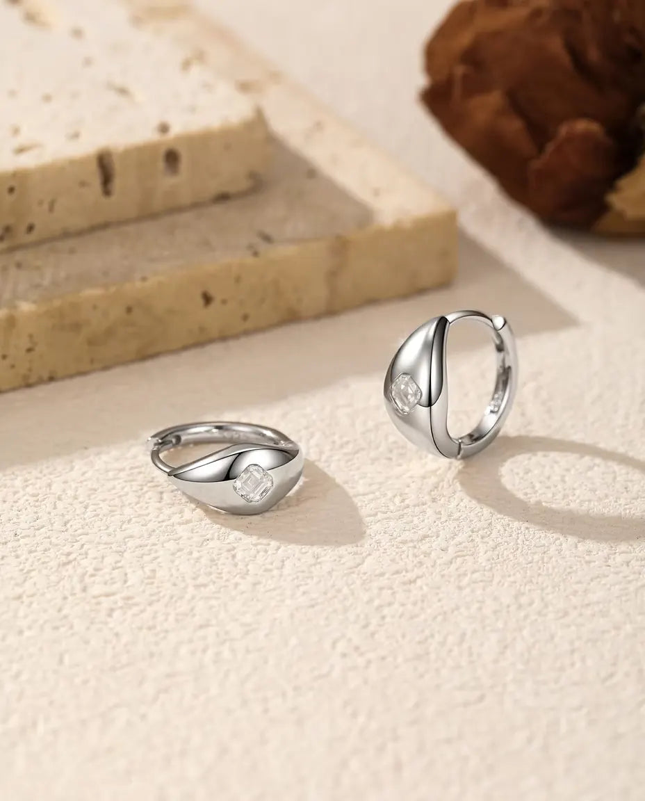 Pendientes de Aro Huggie de Moissanita Talla Asscher en Plata 925 - el par sobre una superficie clara con textura, uno tumbado y otro de pie