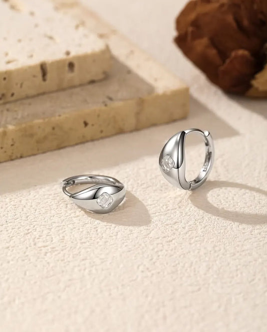 Pendientes de Aro Huggie de Moissanita Talla Asscher en Plata 925 - el par sobre una superficie clara con textura, uno tumbado y otro de pie