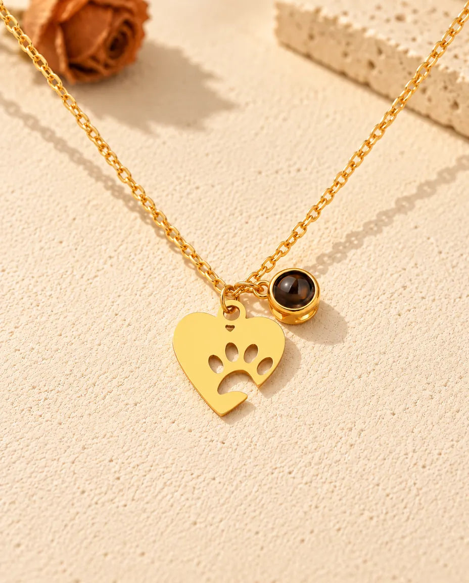 Vista frontal del collar dorado de corazón con huella y su bolita de proyección