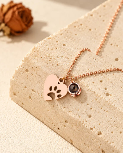 Collar color oro rosa con corazón de huella y bolita de proyección, apoyado en un bloque de travertino