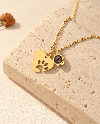 Collar dorado de corazón con huella y bolita de proyección, apoyado sobre travertino