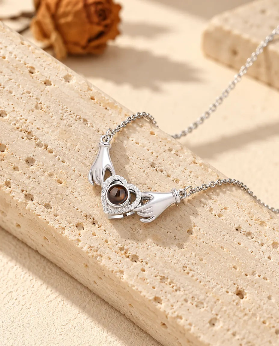 Collar con foto dentro plateado con dos manitas que sujetan un corazón de circonitas, apoyado en un bloque de travertino