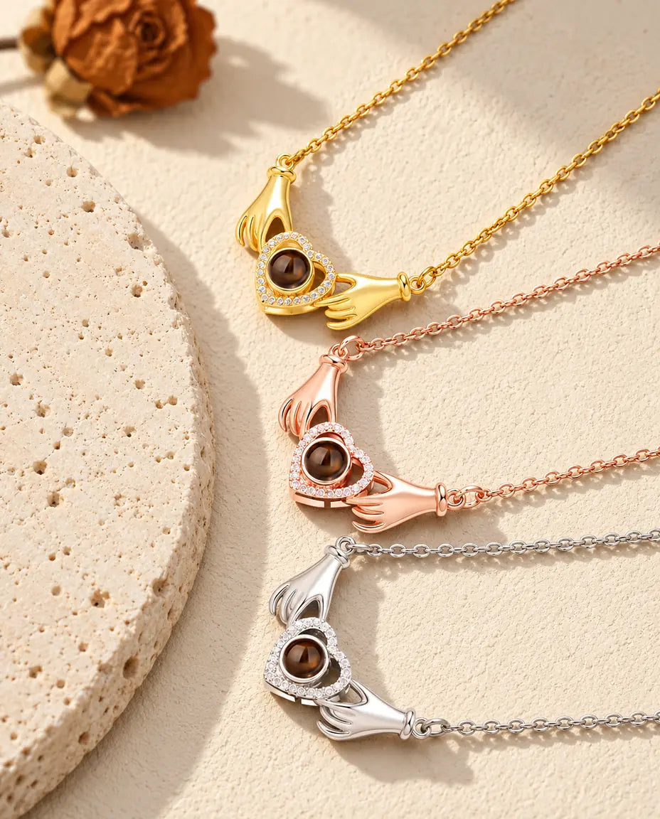 Collares de manos con corazón en dorado, color oro rosa y plateado, apilados junto a una losa de travertino