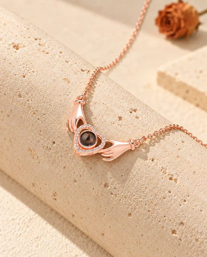 Collar color oro rosa de dos manos con corazón y foto dentro, colgado sobre un bloque de travertino