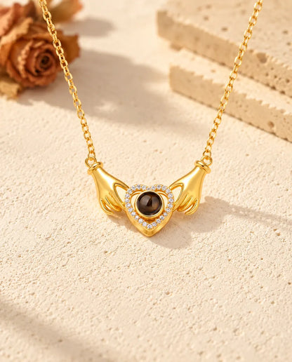 Vista frontal del colgante dorado de manos y corazón, con la cadena enganchada en cada muñeca