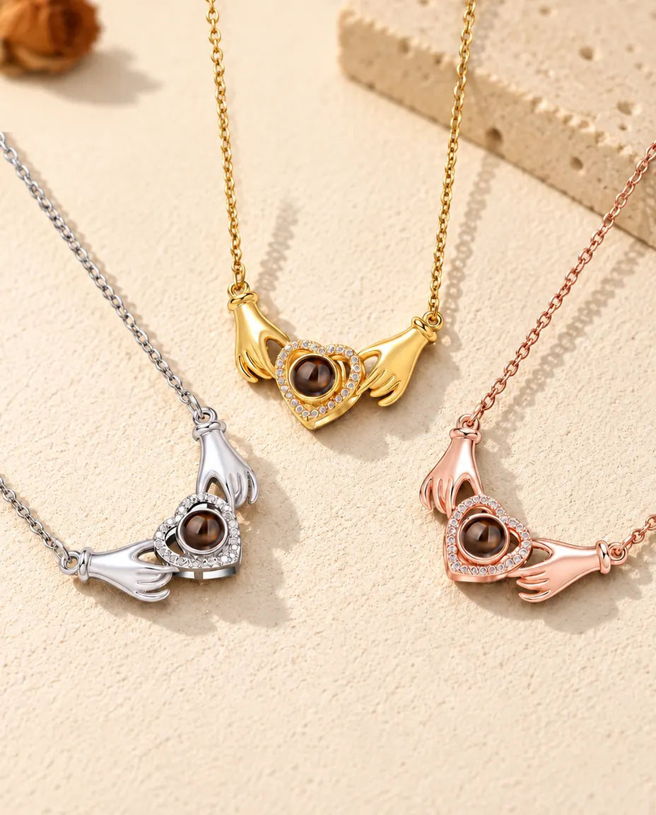 Los tres colores del collar de manos con corazón y foto dentro, en fila sobre yeso color crema