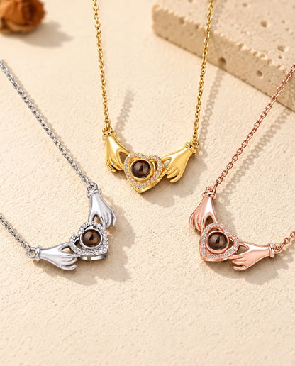 Los tres colores del collar de manos con corazón y foto dentro, en fila sobre yeso color crema