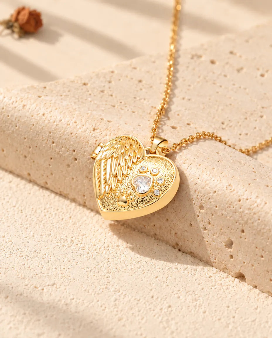 Collar relicario de corazón dorado con ala de ángel y huella apoyado sobre un bloque de travertino