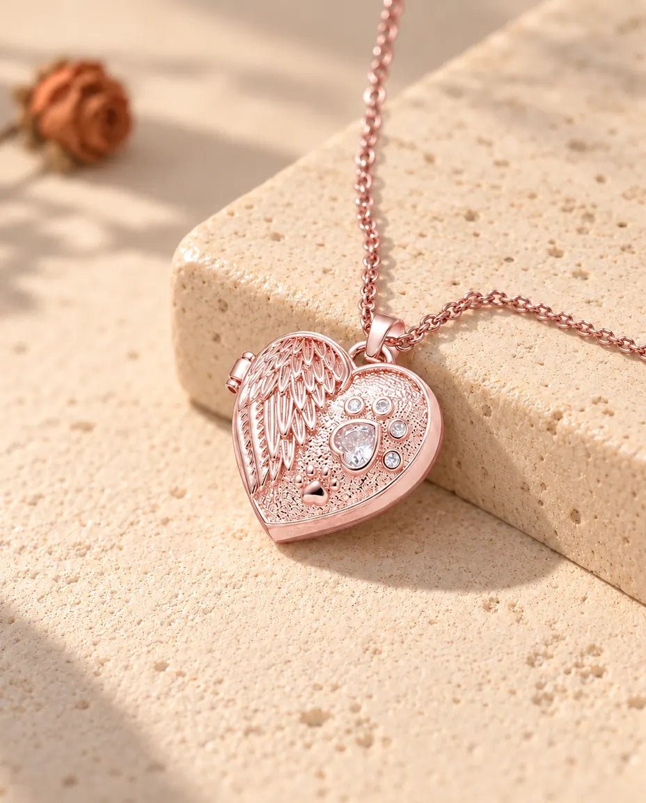 Collar relicario color oro rosa con ala de ángel y huella sobre travertino con luz cálida de sol