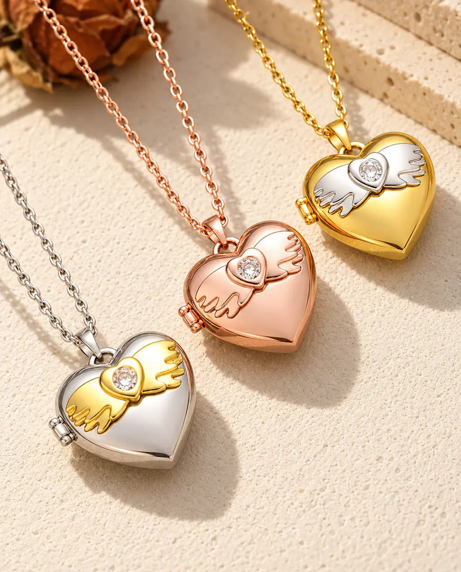 Relicarios de corazón con alas de ángel en plateado, color oro rosa y dorado, colocados en diagonal sobre yeso crema