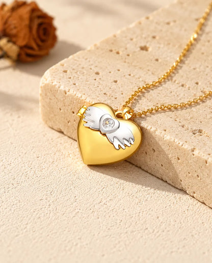 Collar relicario de corazón dorado con las alas plateadas, apoyado en un bloque de travertino