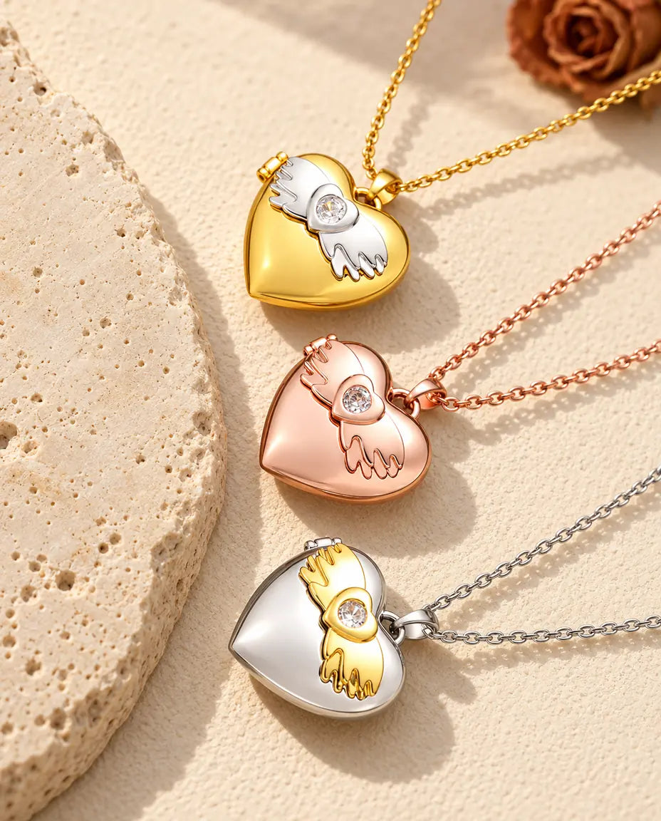 Relicarios de corazón con alas de ángel dorado, color oro rosa y plateado apilados junto a una losa de travertino