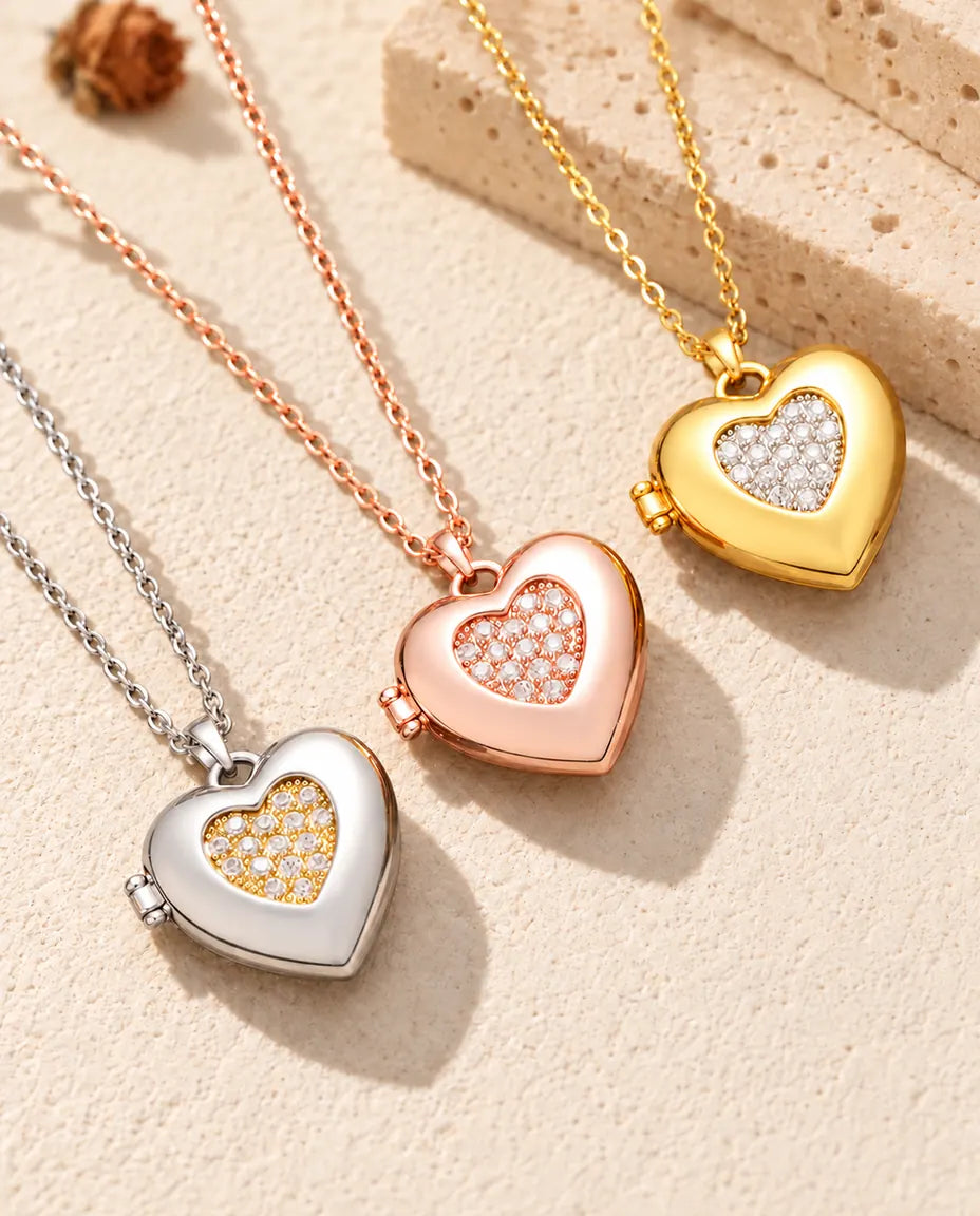 Relicarios de corazón con centro de circonitas en plateado, color oro rosa y dorado, colocados en diagonal sobre yeso crema