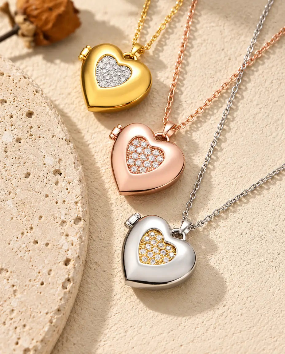 Relicarios de corazón con centro de circonitas dorado, color oro rosa y plateado apilados junto a una losa de travertino