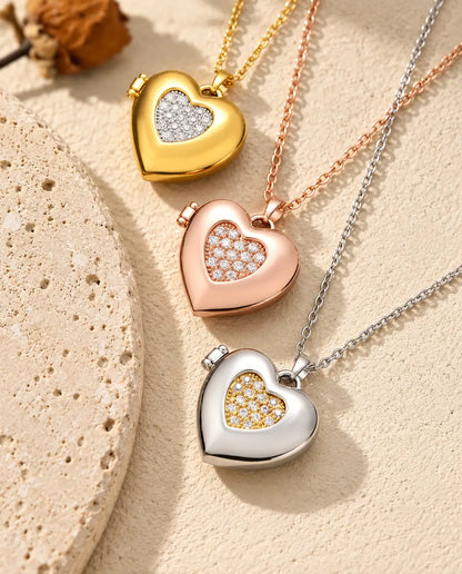 Relicarios de corazón con centro de circonitas dorado, color oro rosa y plateado apilados junto a una losa de travertino