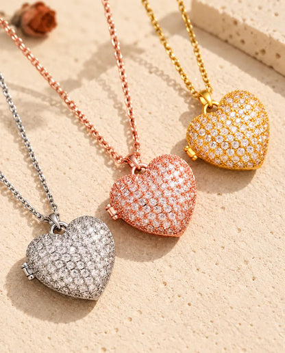 Relicarios de corazón cuajados de circonitas en plateado, color oro rosa y dorado, colocados en diagonal sobre yeso crema