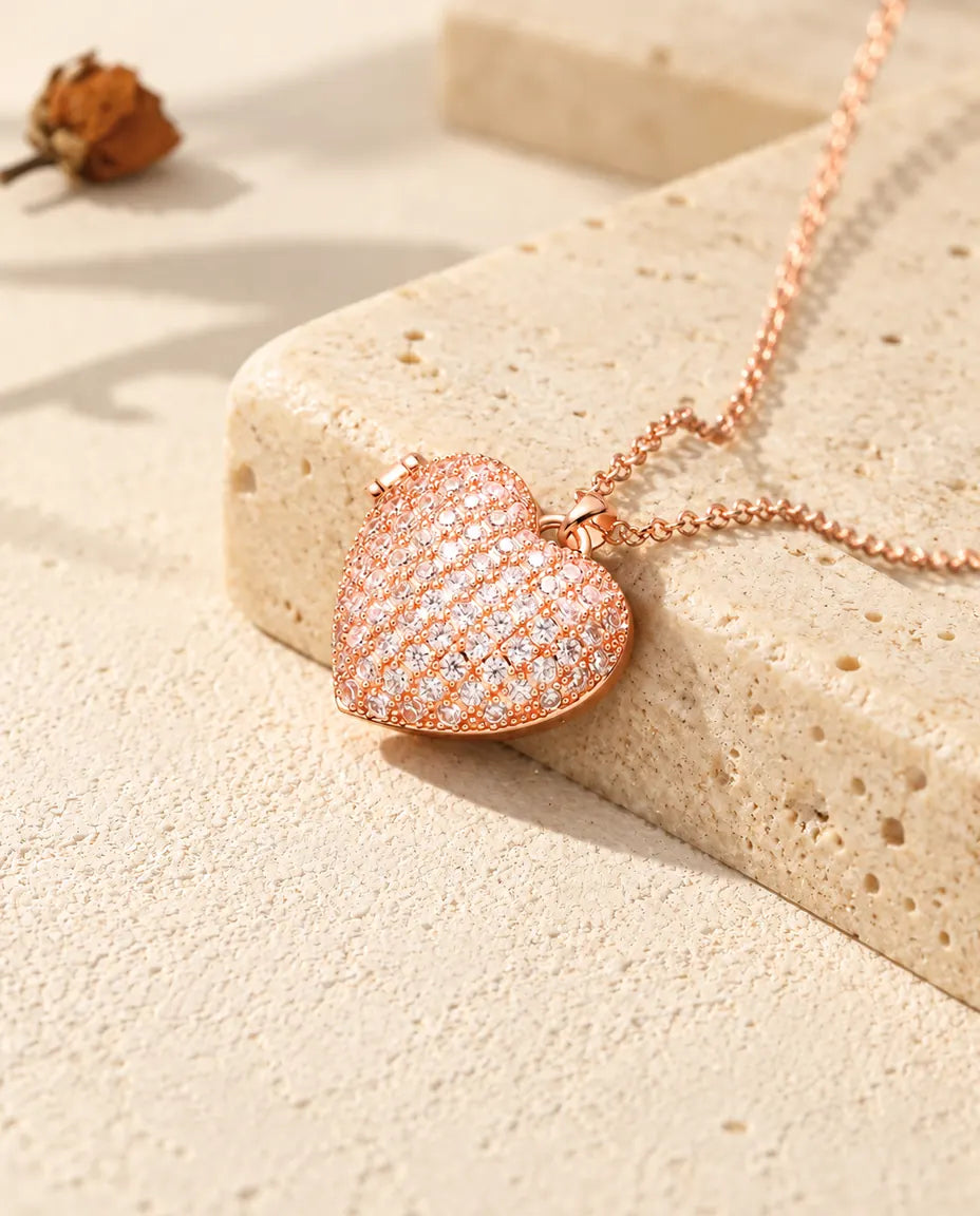 Collar relicario de corazón cuajado de circonitas en color oro rosa sobre travertino, con luz cálida de sol