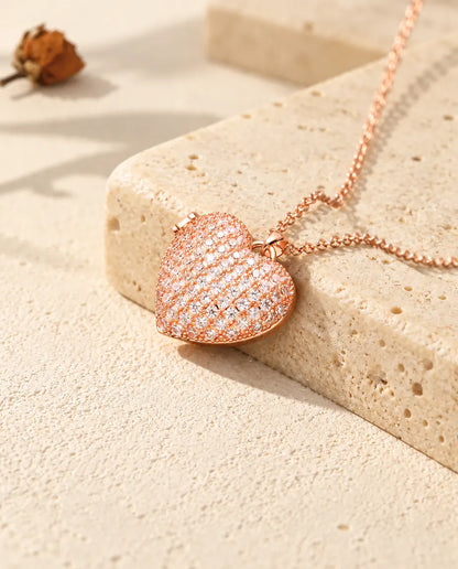Collar relicario de corazón cuajado de circonitas en color oro rosa sobre travertino, con luz cálida de sol