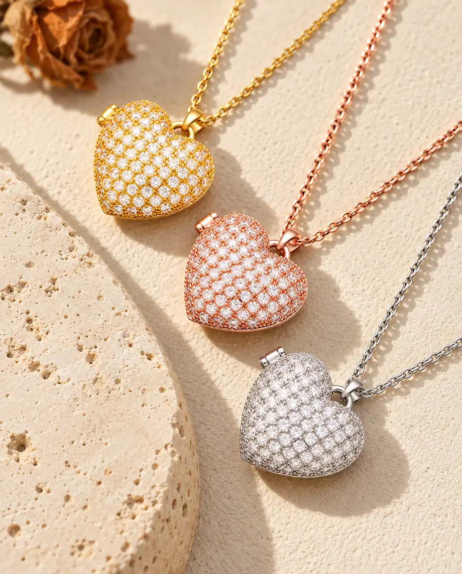 Relicarios de corazón cuajados de circonitas dorado, color oro rosa y plateado apilados junto a una losa de travertino