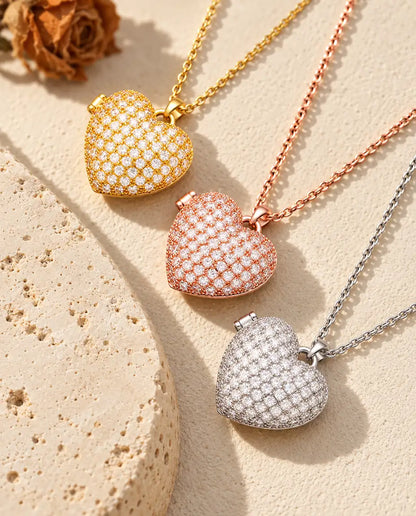 Relicarios de corazón cuajados de circonitas dorado, color oro rosa y plateado apilados junto a una losa de travertino