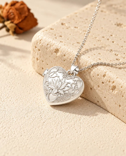 Relicario con foto de corazón plateado con flor de loto y volutas en relieve, apoyado sobre un bloque de travertino