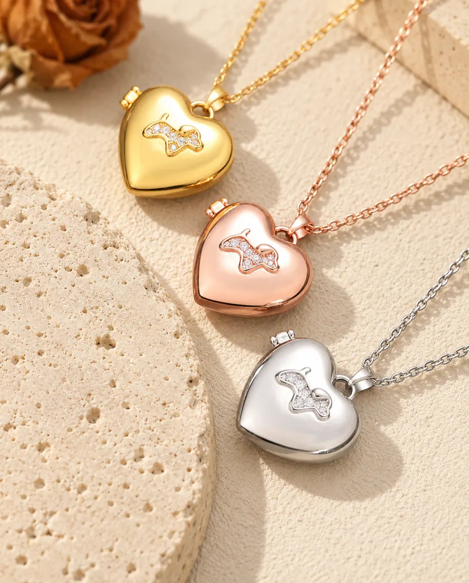 Relicarios de corazón con perrito dorado, color oro rosa y plateado apilados junto a una losa de travertino