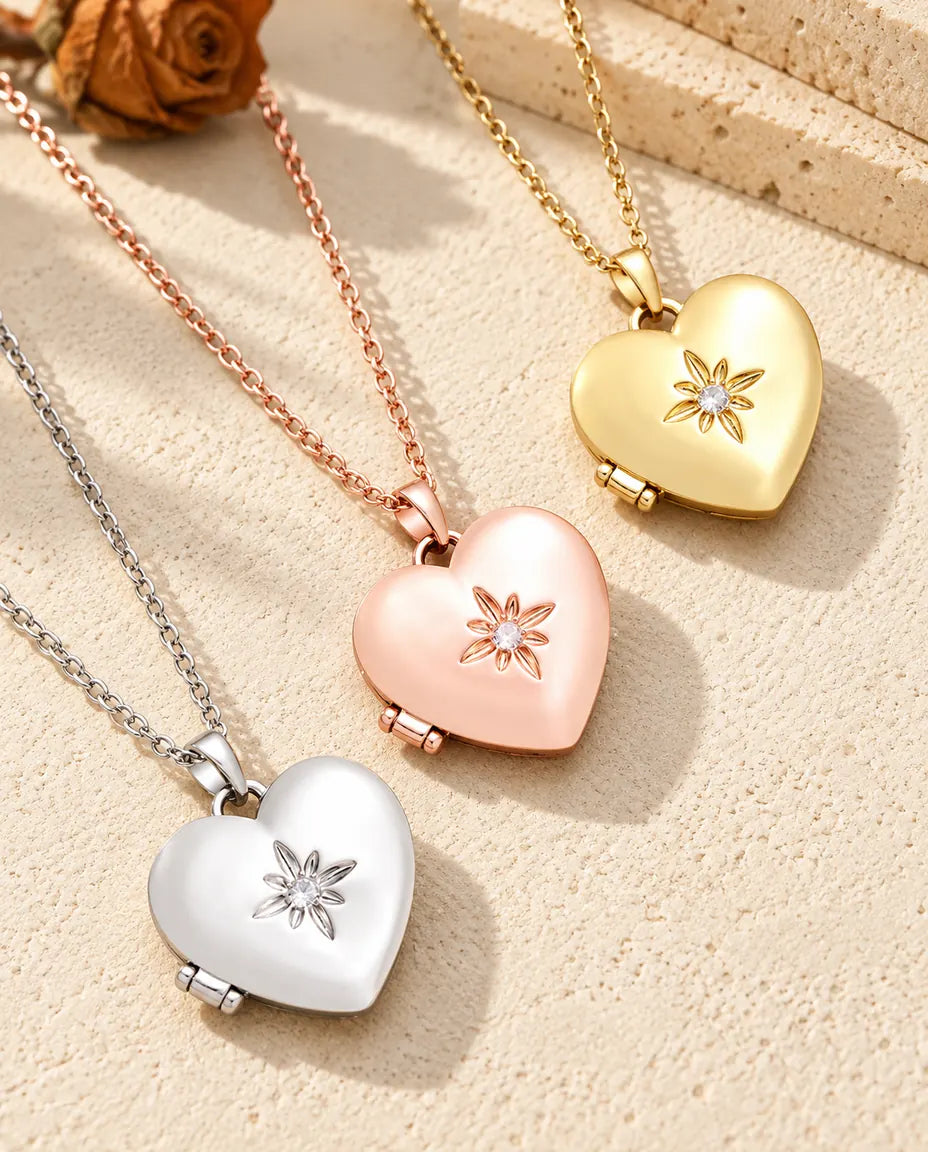 Relicarios de corazón satinados con flor en plateado, color oro rosa y dorado, colocados en diagonal sobre yeso crema