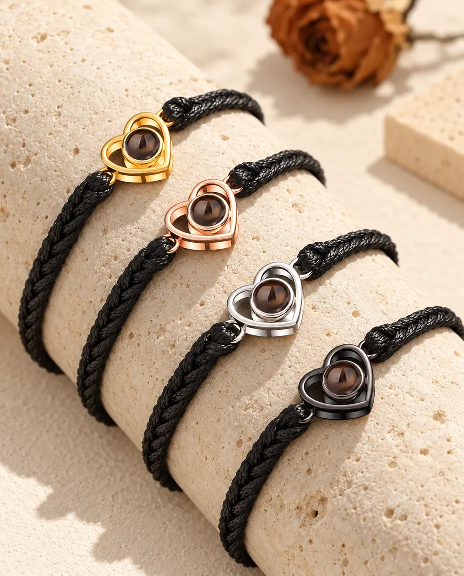 Cuatro pulseras de cordón trenzado negro con corazón abierto en cada color sobre un bloque de travertino