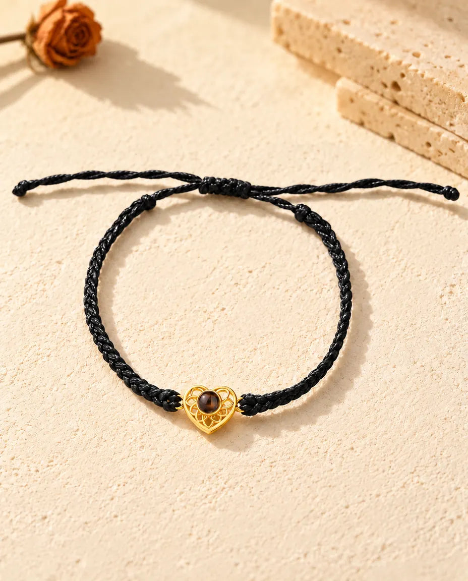 Vista cenital de la pulsera de corazón dorado en cordón trenzado fino negro, con el nudo corredizo y sus extremos