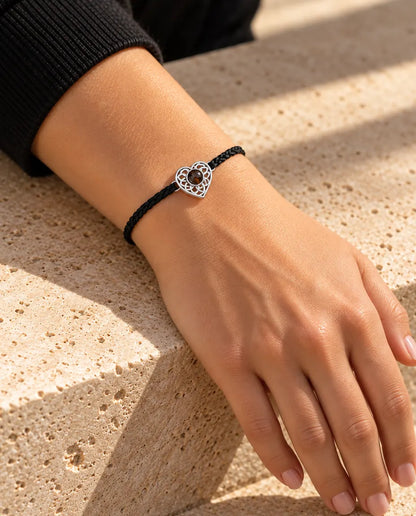 Mujer con la pulsera de cordón trenzado fino negro y corazón plateado en la muñeca, bajo una cálida luz de sol