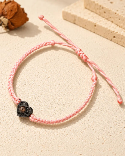 Pulsera de cordón trenzado fino rosa con corazón negro que sujeta una pequeña piedra marrón de proyección