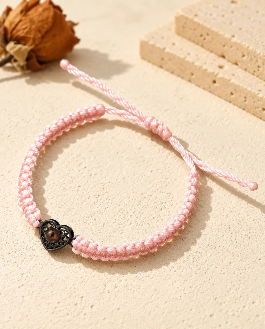 Pulsera de cordón trenzado rosa con corazón negro que sujeta una pequeña piedra marrón de proyección