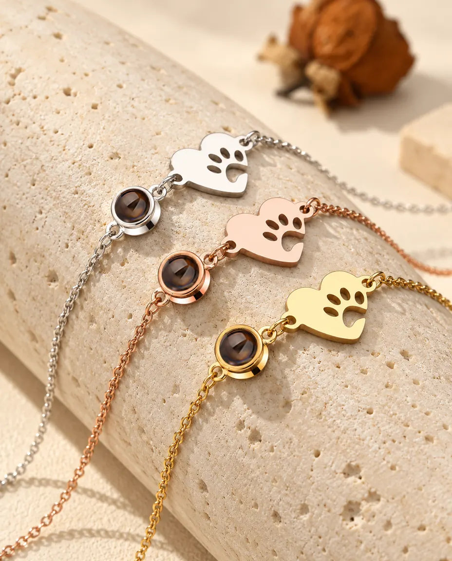 Tres pulseras de corazón con huella de pata en plateado, color oro rosa y dorado sobre un bloque de travertino