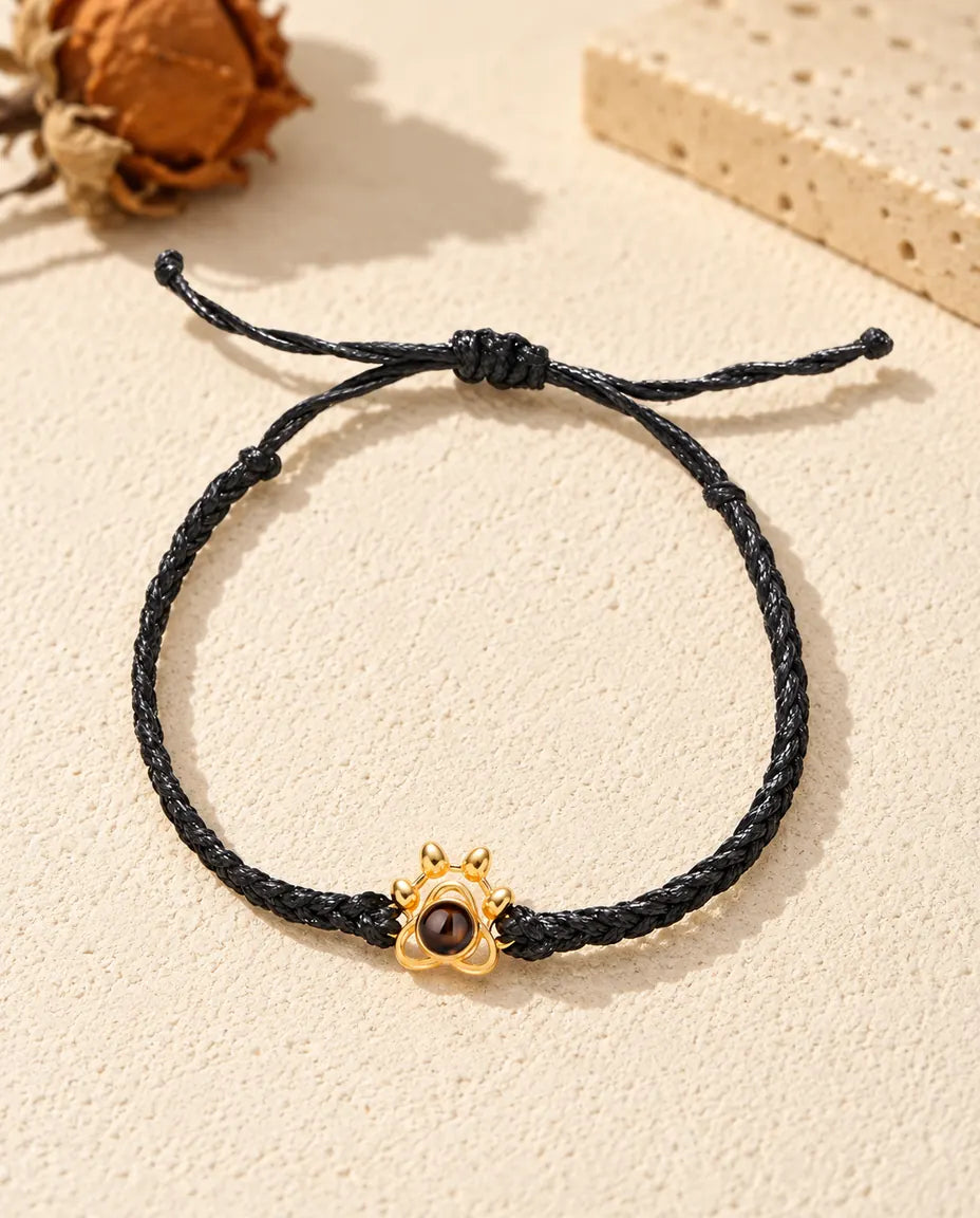 Vista cenital de la pulsera de huella dorada en cordón trenzado negro, con el nudo corredizo y sus extremos