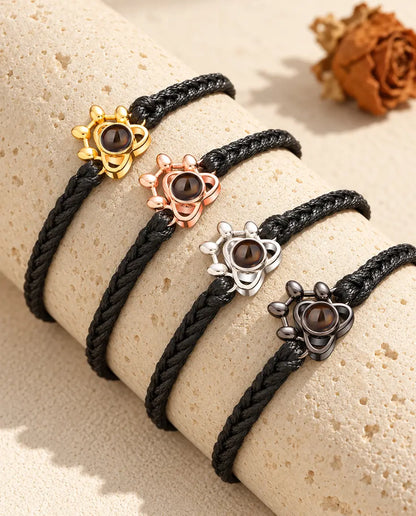 Cuatro pulseras de cordón trenzado negro con colgante huella en cada color sobre un bloque de travertino