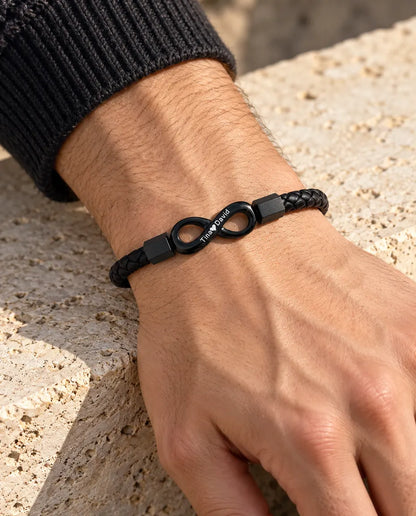 Hombre con la pulsera de cuero sintético trenzado e infinito negro grabado en la muñeca, junto a un jersey de punto gris antracita