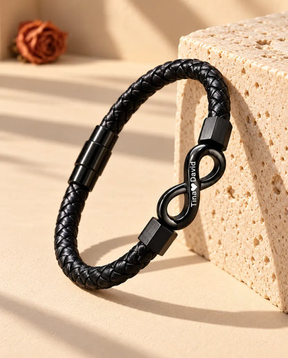 Pulsera de cuero sintético trenzado negro con infinito grabado, apoyada de pie contra un bloque de travertino